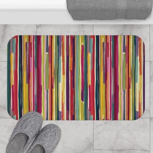 Puede incluir: Una alfombra de baño rectangular con un diseño abstracto vibrante. Rayas verticales en varios colores, incluyendo rojo, amarillo, verde y morado, crean un patrón animado. La alfombra está colocada sobre un suelo embaldosado, con un par de zapatillas grises.