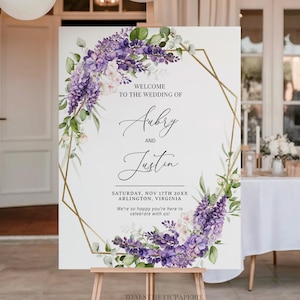 May include: A wedding welcome sign with the names "Aubry and Justin" displayed within a gold geometric frame, adorned with purple and white floral accents. The sign is on a wooden easel. The text reads "Welcome to the wedding of" and the wedding date and location.