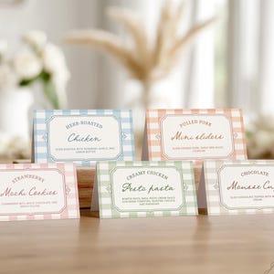 May include: A collection of six food labels with a gingham pattern in pastel colors. The labels display menu items such as "Herb-Roasted Chicken" and "Strawberry Mochi Cookies". The labels are arranged on a wooden surface.