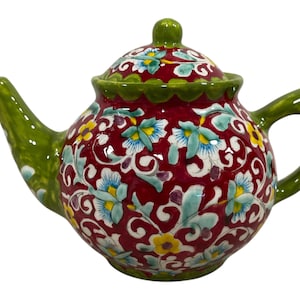 Peut inclure: Une théière en céramique rouge et verte avec un motif floral. La théière a un couvercle et un bec verseur. Le motif floral est dans des tons de bleu, jaune et vert.