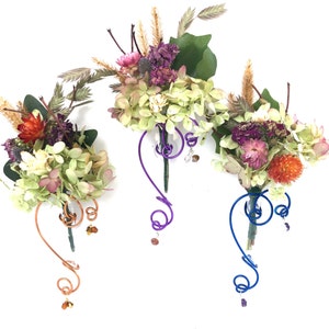 May include: Three dried flower arrangements in small glass vases with wire stems. The stems are decorated with colorful beads. The vases are attached to a wire that is shaped like a question mark. The flowers are a mix of white, pink, purple, and orange.