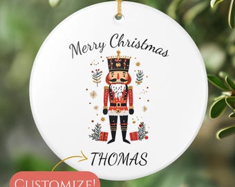 May include: White ceramic Christmas ornament with a gold string. The ornament features a nutcracker illustration with the text "Merry Christmas" and the name "THOMAS". A red "Customize!" button is in the lower left.