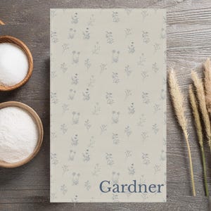 Pode incluir: Um pano de cozinha retangular bege com um delicado padrão floral em azul claro. A palavra "Gardner" está impressa em uma fonte azul escura na parte inferior. Duas tigelas de madeira cheias de pó branco estão à esquerda.