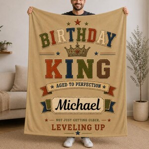 May include: A tan fleece blanket featuring the text BIRTHDAY KING in multi-colored block letters, a crown graphic, and the name Michael. Additional text includes AGED TO PERFECTION, NOT JUST GETTING OLDER, and LEVELING UP with star accents.
