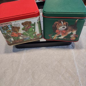 May include: Two vintage metal tins. One is red with a bear scene, the other is green with a puppy illustration. Both have square shapes and lids. The bear tin shows bears in a kitchen setting. The puppy tin has a puppy with a red bow.