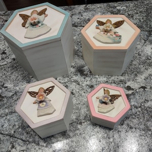 Puede incluir: Cuatro cajas de madera hexagonales con figuritas de ángeles en las tapas. Las cajas están pintadas en varios colores pastel, incluyendo rosa, azul y melocotón. Los ángeles sostienen flores y tienen alas.