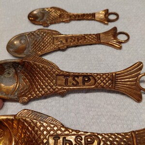 May include: A set of antique brass measuring spoons and a ring, all in a fish shape. The spoons are labeled with "TSP" and "Tbsp" measurements. The spoons have a textured scale pattern and a loop at the end for hanging.