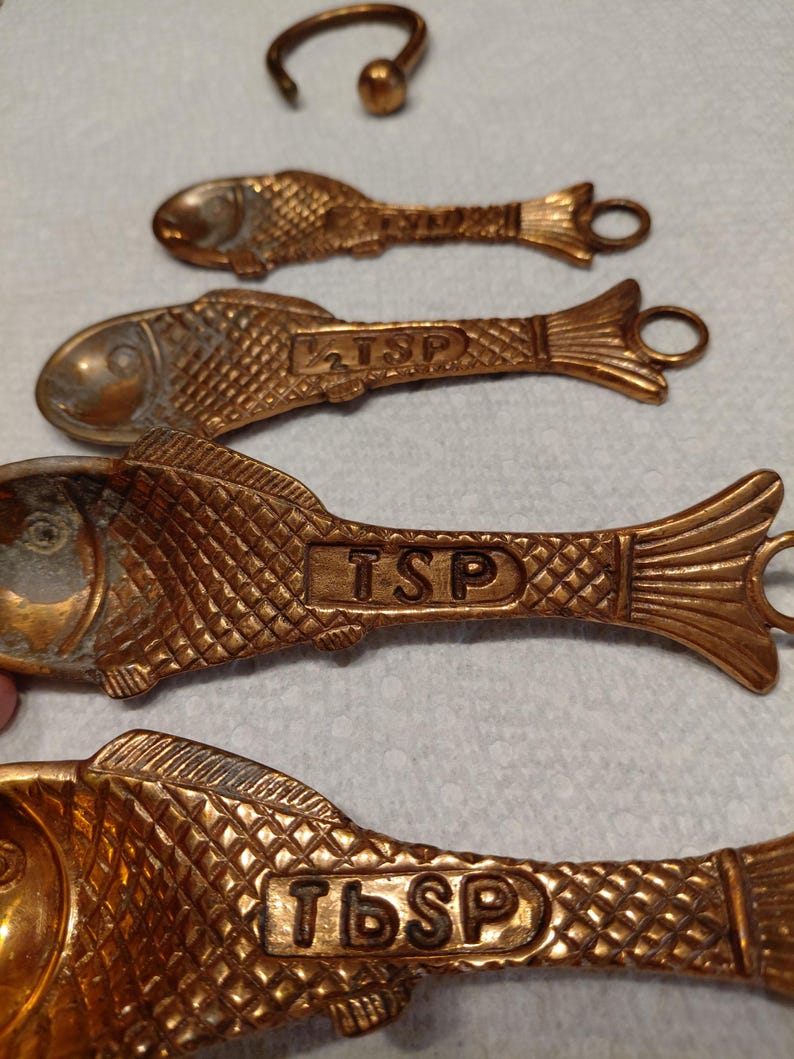 May include: A set of antique brass measuring spoons and a ring, all in a fish shape. The spoons are labeled with "TSP" and "Tbsp" measurements. The spoons have a textured scale pattern and a loop at the end for hanging.