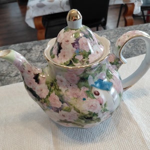 Könnte beinhalten: Dekorative Teekanne mit floralem Muster, verziert mit rosa, lila und grünen Blumen. Sie hat einen goldfarbenen Griff und Akzente, einen hellblauen Ausguss und Henkel sowie einen goldenen Knauf auf dem Deckel.