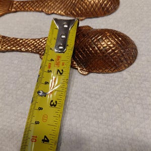 May include: Bronze-colored fish-shaped spoons with textured scales. A yellow measuring tape shows the spoon is approximately 4 inches long. The spoons are arranged on a white surface.