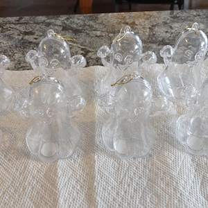 Könnte beinhalten: Klare Engel-Ornamente aus Kunststoff mit goldfarbenen Aufhängeschlaufen. Die Ornamente sind auf einem weißen Papiertuch angeordnet. Die Engel haben ausgestreckte Arme und detaillierte Gesichtszüge.