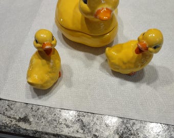 May include: Three yellow ceramic duck figurines. The largest duck is a container with a lid. The ducks have black eyes and orange beaks.
