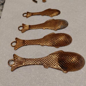 May include: Four decorative, fish-shaped spoons and a small ring, all in a metallic gold color. The spoons have a textured scale pattern and a loop at the end of the handle. The ring is curved with a small ball at the end.