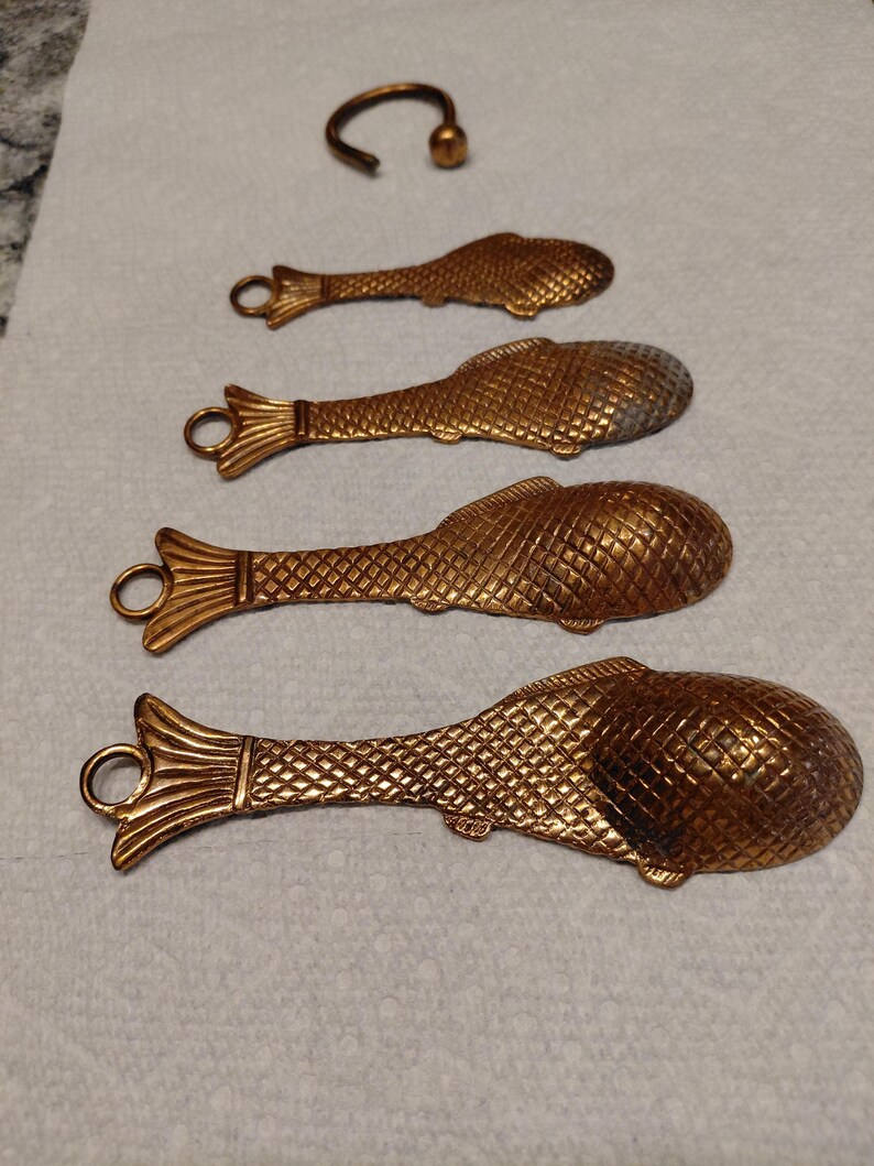 May include: Four decorative, fish-shaped spoons and a small ring, all in a metallic gold color. The spoons have a textured scale pattern and a loop at the end of the handle. The ring is curved with a small ball at the end.