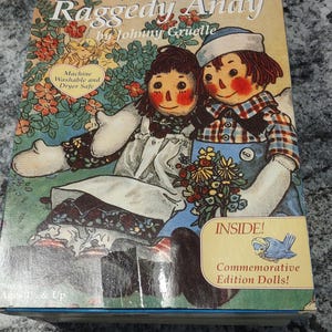 May include: A book cover featuring Raggedy Ann and Raggedy Andy, two iconic rag dolls, sitting together in a garden setting. The book title is "The Adventures of Raggedy Ann and Raggedy Andy" by Johnny Gruelle. The cover also states "Machine Washable and Dryer Safe" and "Inside! Commemorative Edition Dolls!"