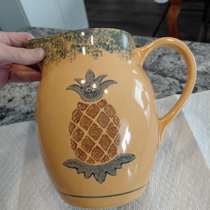 May include: A yellow ceramic pitcher featuring a brown pineapple design with a blue-grey crown and base. The rim and handle are dark green. The pitcher is suitable for serving drinks.