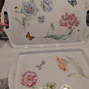 May include: Two rectangular serving trays with floral and butterfly designs. The trays are white with handles and feature blue, pink, and orange flowers, along with butterflies and dragonflies. The trays have a glossy finish.
