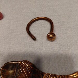 May include: A bronze-colored key ring with a rounded ball at the end of the curved handle. A decorative fish-shaped spoon with the text "VATS SP" is also visible. The items are on a light-colored surface.