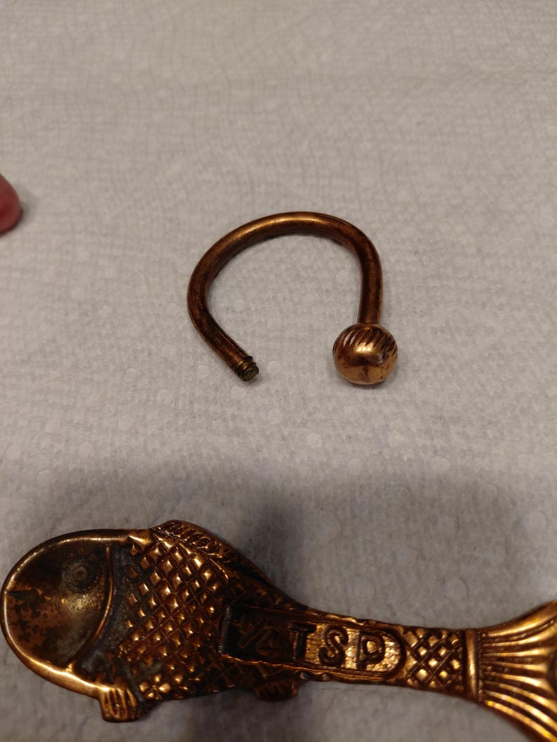 May include: A bronze-colored key ring with a rounded ball at the end of the curved handle. A decorative fish-shaped spoon with the text "VATS SP" is also visible. The items are on a light-colored surface.
