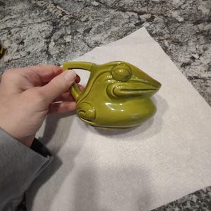 May include: A green ceramic frog pitcher with a handle. The frog has a detailed face with eyes and a wide mouth.