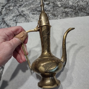 Puede incluir: Una tetera de latón de color dorado con un diseño grabado decorativo. La tetera tiene una tapa con bisagras y un pico con un asa curva.