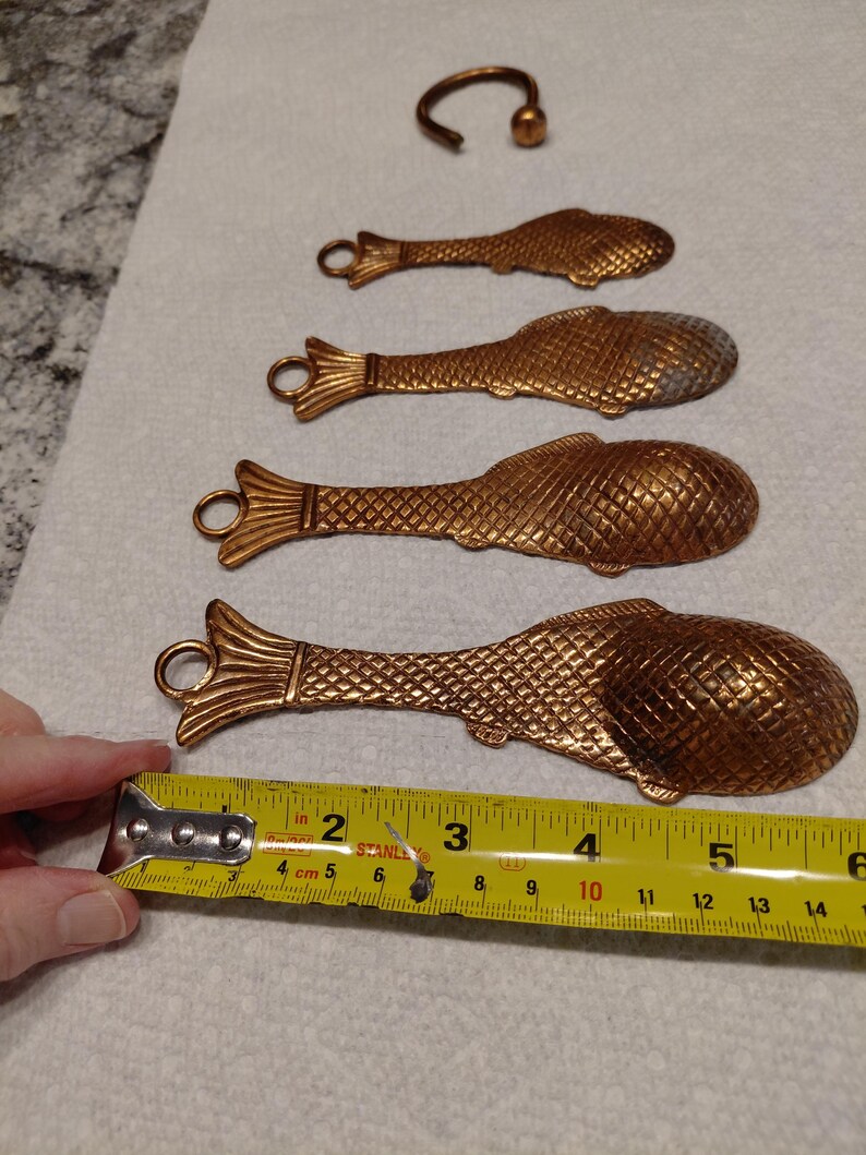 May include: Four bronze-colored fish-shaped spoons and a small hook. The spoons are approximately 5 inches long. The spoons have a textured scale pattern and a loop at the tail end. The hook has a small ball at the end.