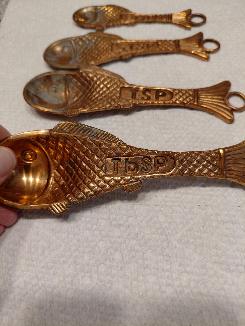 May include: Four gold-colored measuring spoons shaped like fish. The spoons have a textured scale pattern and the text "TSP" or "TBSP" is embossed on the body. Each spoon has a loop at the end of the handle.
