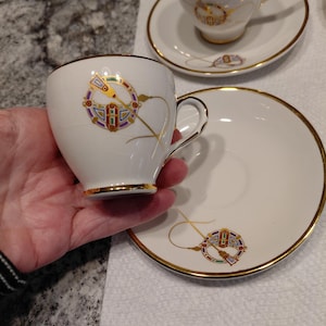 May include: White teacups and saucers with gold trim and a colorful circular design. The teacups have a small handle and the saucers are round. The design includes purple, red, green, and yellow accents.