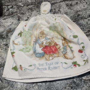 May include: White cotton tea towel with a crocheted loop. The towel features a colorful illustration of Peter Rabbit and his family from the book "The Tale of Peter Rabbit".