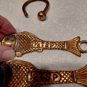 May include: Two bronze-colored fish-shaped measuring spoons, one marked "1/4 TSP" and the other "1/2 TSP". A bronze ring with a ball-shaped end is also visible. The spoons have a textured, scale-like pattern.