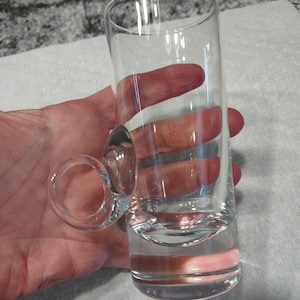 Puede incluir: Un vaso de chupito de vidrio transparente con una pequeña asa. El vaso es cilíndrico con un fondo redondeado.