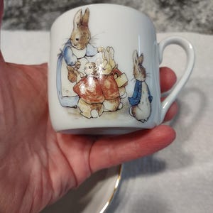 Può includere: Una tazza da tè in porcellana bianca con manico e un'illustrazione colorata di Peter Rabbit e la sua famiglia. La tazza ha un bordo dorato.