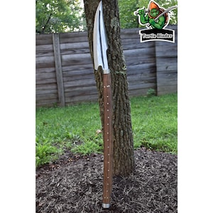 May include: A long, silver spear with a wooden handle. The handle is decorated with small, silver studs. The spear is leaning against a tree trunk.