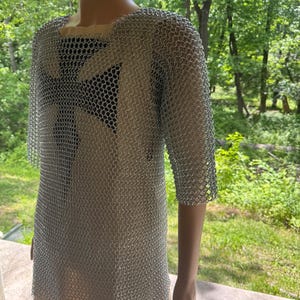 May include: A silver chainmail shirt with short sleeves and a square neckline. A black and white cross design is visible through the chainmail. The shirt is displayed on a mannequin against a backdrop of green trees.