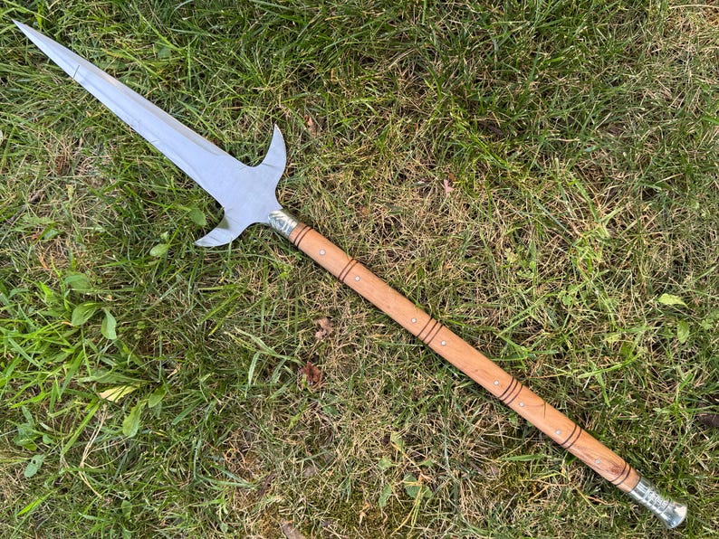 Pu&ograve; includere: Un'asta con una lama in acciaio lucido e un'asta in legno. La lama ha una punta centrale e due sporgenze laterali curve. L'asta &egrave; marrone chiaro con fasce e rivetti metallici decorativi. L'asta &egrave; appoggiata sull'erba verde.