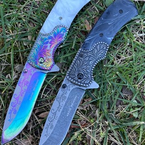 Op de afbeelding: Twee inklapbare messen met florale gravures op de bladen en kropstukken. Het linker mes heeft een wit handvat en een regenboogkleurig iriserend blad. Het rechter mes heeft een zwart gemarmerd handvat en een donkergrijs blad met de tekst Old River.