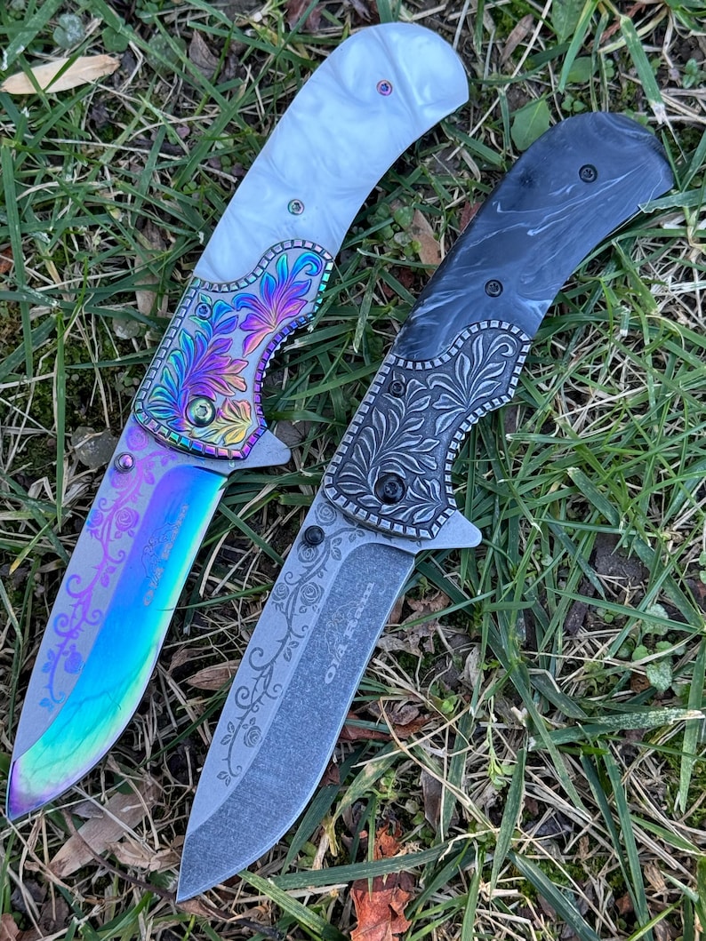 Op de afbeelding: Twee inklapbare messen met florale gravures op de bladen en kropstukken. Het linker mes heeft een wit handvat en een regenboogkleurig iriserend blad. Het rechter mes heeft een zwart gemarmerd handvat en een donkergrijs blad met de tekst Old River.