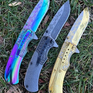 Op de afbeelding: Drie inklapbare messen met florale gravures op de bladen en handvatten. Het linker mes heeft een iriserende regenboogafwerking, het middelste mes is matgrijs met een zwarte clip en het rechter mes heeft een gepolijste goudkleurige afwerking.