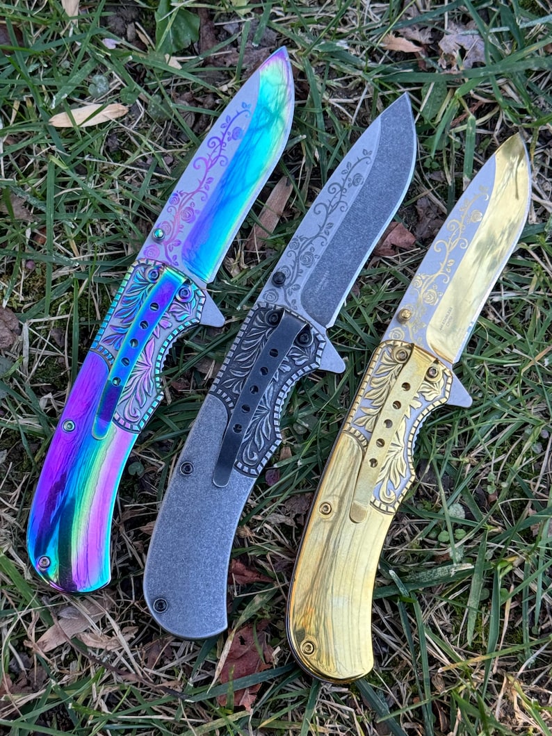 Op de afbeelding: Drie inklapbare messen met florale gravures op de bladen en handvatten. Het linker mes heeft een iriserende regenboogafwerking, het middelste mes is matgrijs met een zwarte clip en het rechter mes heeft een gepolijste goudkleurige afwerking.