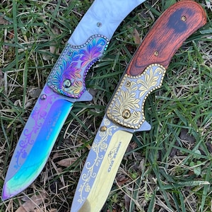 Op de afbeelding: Twee inklapbare messen met versierde metalen kropstukken en bewerkte bladen. Het linker mes heeft een wit handvat en een regenboogkleurig blad met florale gravures. Het rechter mes heeft een handvat met houtnerf en een goudkleurig blad met rozenmotieven.