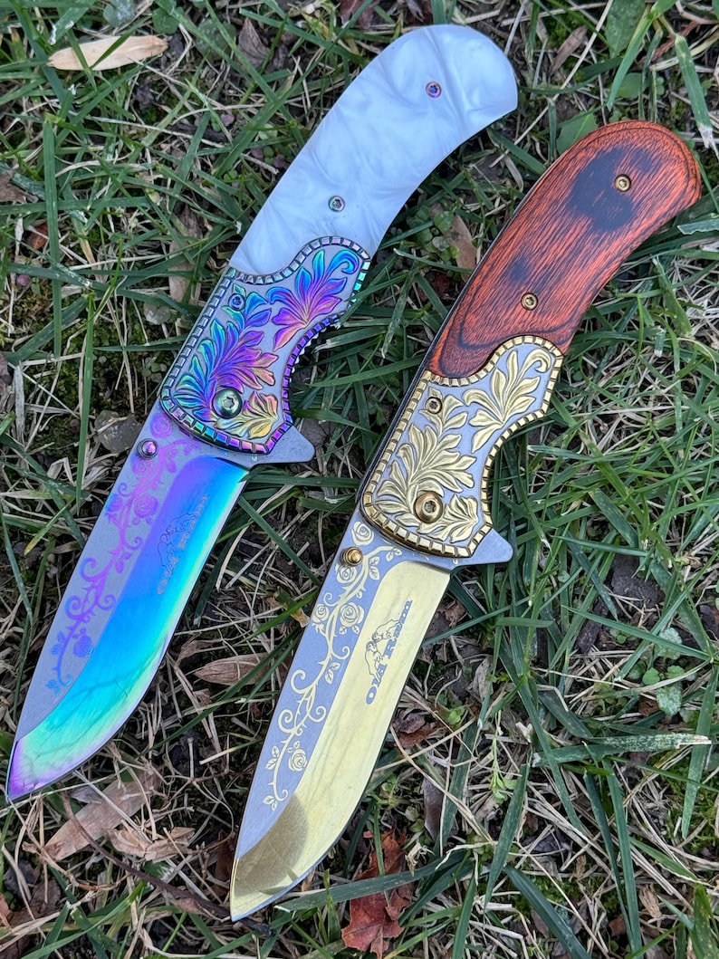 Op de afbeelding: Twee inklapbare messen met versierde metalen kropstukken en bewerkte bladen. Het linker mes heeft een wit handvat en een regenboogkleurig blad met florale gravures. Het rechter mes heeft een handvat met houtnerf en een goudkleurig blad met rozenmotieven.