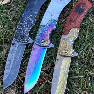 Op de afbeelding: Drie inklapbare messen met gedetailleerde bloemgravures op de bladen en kropstukken. Het linker mes heeft een zwart handvat, het middelste mes heeft een wit handvat met een iriserend regenboogblad en het rechter mes heeft een houtlook handvat.