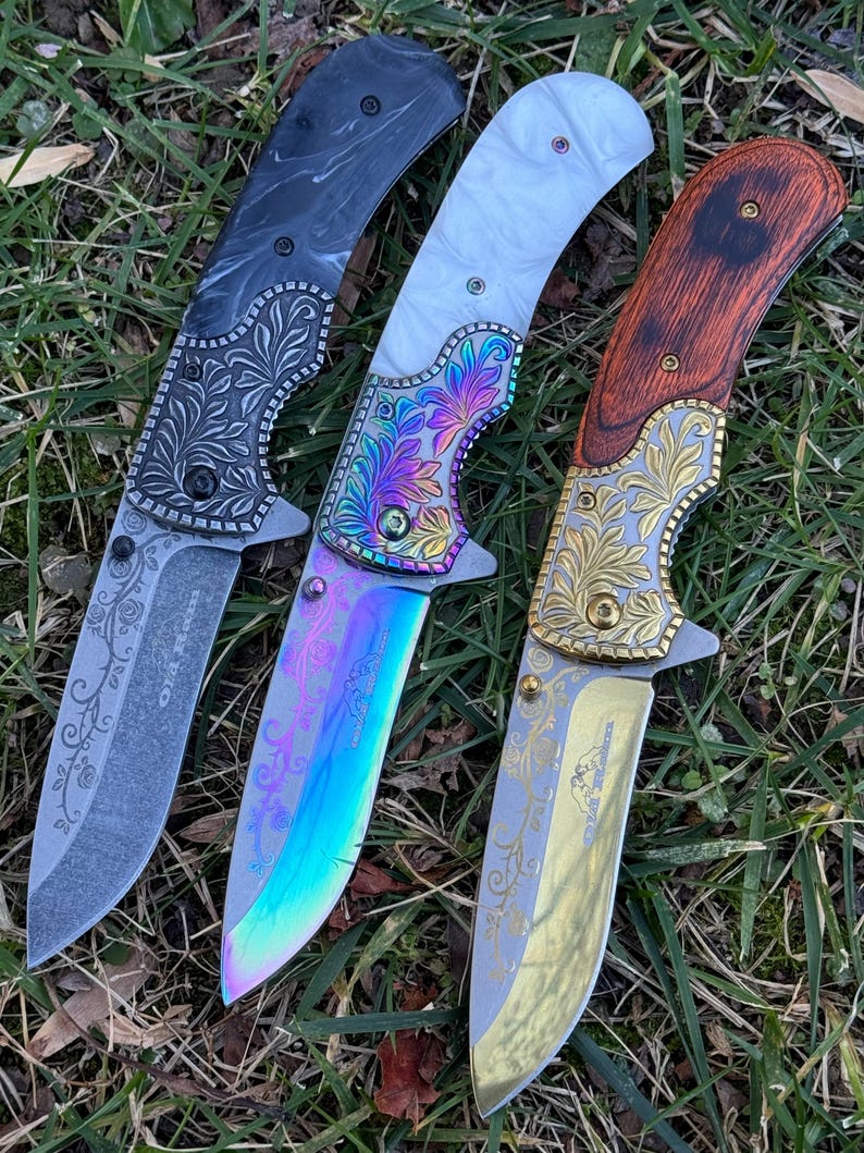 Op de afbeelding: Drie inklapbare messen met gedetailleerde bloemgravures op de bladen en kropstukken. Het linker mes heeft een zwart handvat, het middelste mes heeft een wit handvat met een iriserend regenboogblad en het rechter mes heeft een houtlook handvat.