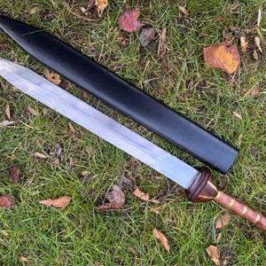 May include: A Roman gladius sword with a wooden handle and a silver blade, resting on a bed of green grass and fallen leaves. The sword is accompanied by a black leather scabbard.