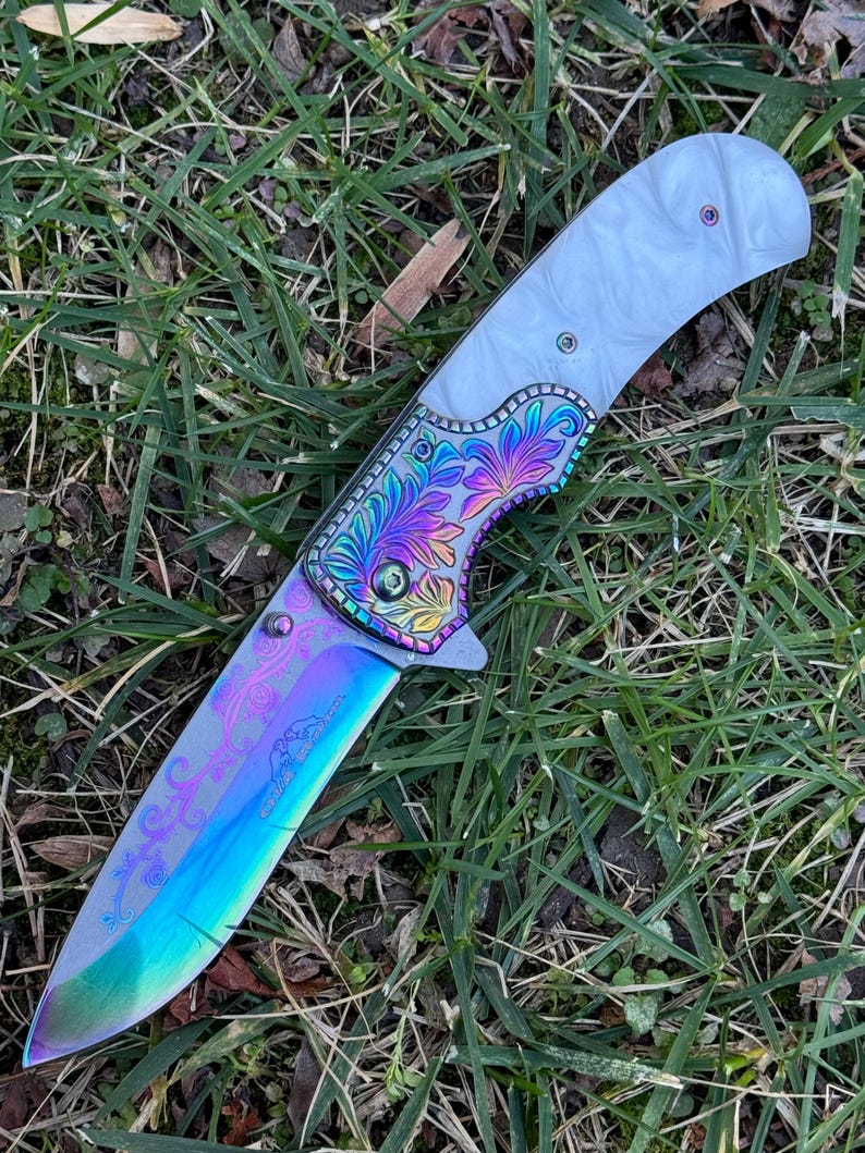 Op de afbeelding: Een vouwmes met een iriserend, regenboogkleurig lemmet voorzien van gegraveerde bloemmotieven en het silhouet van een klein dier. Het handvat heeft een witte, parelmoerachtige greep en een metalen bolster versierd met kleurrijke bloemgravures.