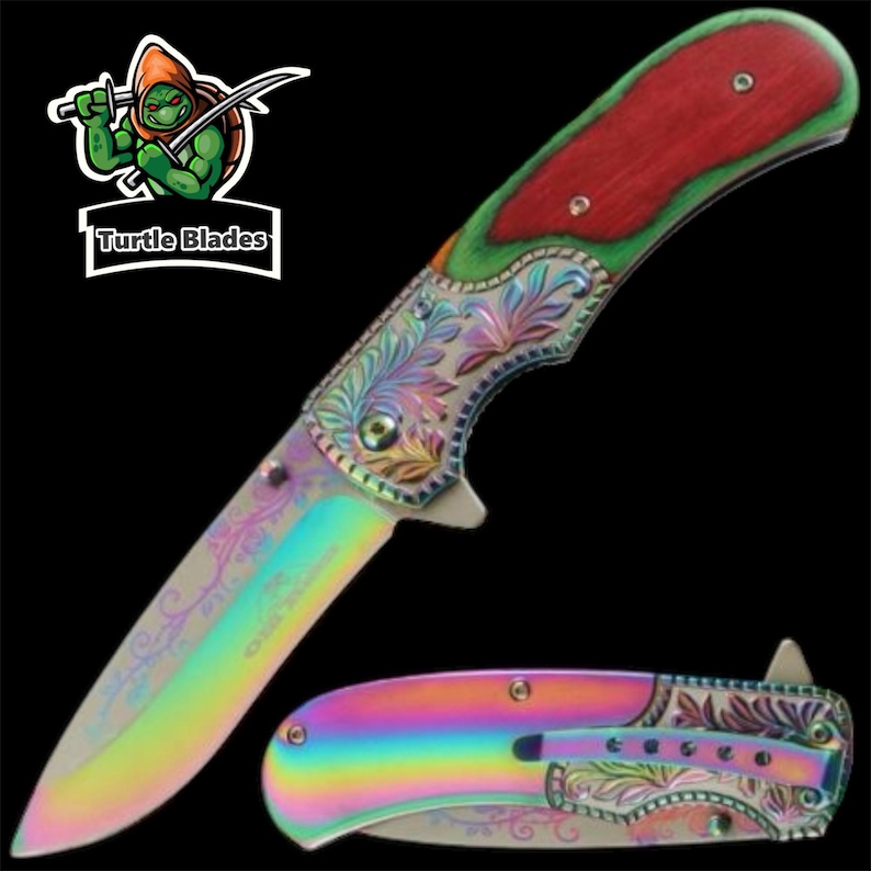 Op de afbeelding: Een vouwmes met een iriserend regenboogkleurig lemmet en handvat met ingewikkelde florale gravures. Het handvat bevat een inleg van rood hout en een metalen zakclip. Een logo met een groene schildpad en de tekst Turtle Blades is zichtbaar.