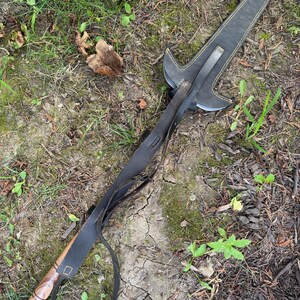 Pu&ograve; includere: Un'asta fatta a mano con manico in legno e lama in metallo. L'asta &egrave; racchiusa in un fodero di pelle nera con cuciture gialle. Un cinturino in pelle nera &egrave; attaccato al fodero e al manico. L'asta &egrave; appoggiata a terra.