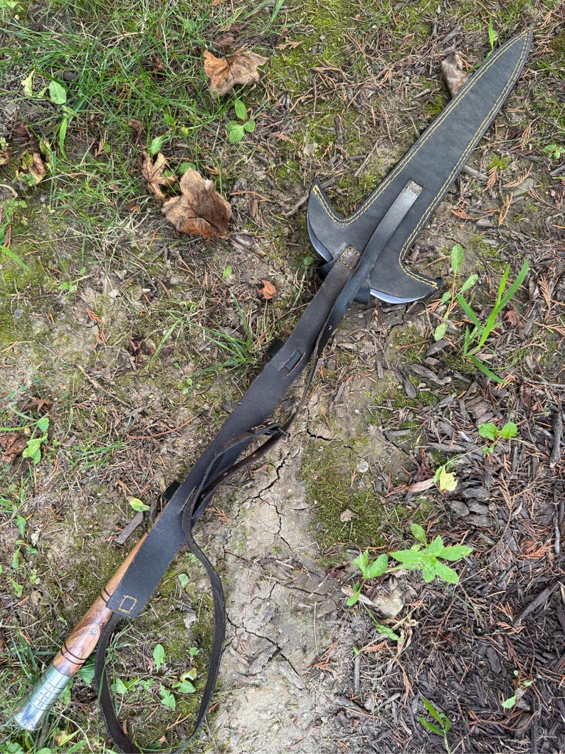 Pu&ograve; includere: Un'asta fatta a mano con manico in legno e lama in metallo. L'asta &egrave; racchiusa in un fodero di pelle nera con cuciture gialle. Un cinturino in pelle nera &egrave; attaccato al fodero e al manico. L'asta &egrave; appoggiata a terra.
