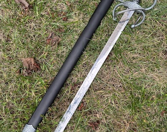 Op de afbeelding: Een decoratief zwaard met een zilveren lemmet en een sierlijke pareerstang, liggend op gras naast de zwarte schede. Het zwaard heeft een gedetailleerd ontwerp op het lemmet en de pareerstang, met een zwarte handgreep. De schede heeft zilveren accenten.