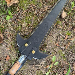 Pu&ograve; includere: Un pugnale fatto a mano con manico in legno e fodero in pelle. La lama &egrave; argentata e il fodero &egrave; nero con cuciture gialle. Il pugnale &egrave; appoggiato su un letto di foglie ed erba.